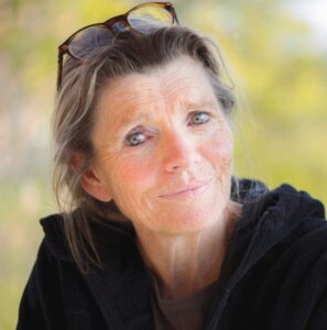 Nahaufnahme einer älteren Frau mit Brille auf dem Kopf, lächelnd im Freien, unscharfer grüner Hintergrund.